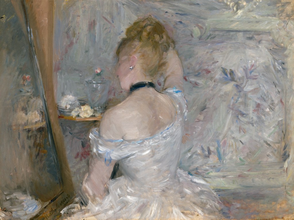 A woman in a white dress with a blue ribbon is seen from the back as she arranges her hair in front of a mirror. The setting has soft, muted colors and floral patterns in the background. Painting by Berthe Morisot.